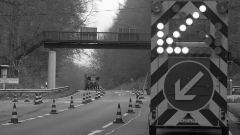 auf der Straße sind Warnkegel aufgestellt, im Vordergrund steht ein Anhänger mit einem leuchtenden Pfeil und ein blaues Verkehrsschild mit weißem Pfeil weisen auf die zu benutzende Fahrbahn